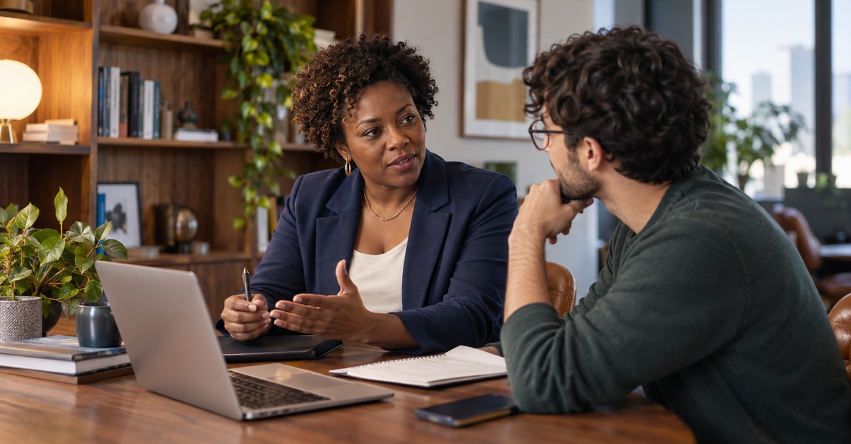 Consultora conversa com um profissional diante do computador e de documentos de trabalho.