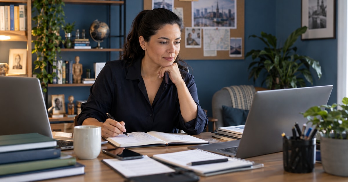 Profissional organiza sua pesquisa com caderno e computadores em uma mesa de trabalho.